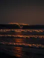 Dark elemental photograph: waves breaking on a black sand beach at ember dusk, the crest of the next swell dissolving into small glowing marks.