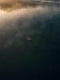 Dawn aerial photograph: a rowboat mid-lake in dawn mist, its wake trailing back toward the dock.
