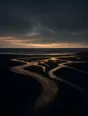 Dark elemental photograph: braided river channels crossing black sand toward a dark sea at dusk, the newest channel in the foreground rendering as glowing amber marks.