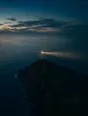 Dawn aerial photograph: a lighthouse on a dark headland at the edge of dawn, its beam still turning.