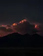 Dark elemental photograph: a towering cloud bank in deep ember light above a black ridge, its highest crown dissolving into glowing marks.