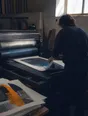 Dark cinematic photograph: A working print shop with a large flatbed press running, one printer's back as they pull a finished sheet clear, printed sheets stacking unevenly beside the machine, hard daylight from a high window.