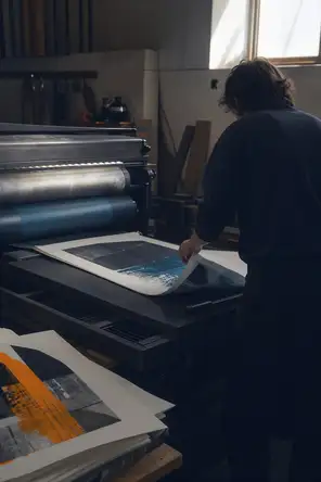 Dark cinematic photograph: A working print shop with a large flatbed press running, one printer's back as they pull a finished sheet clear, printed sheets stacking unevenly beside the machine, hard daylight from a high window.