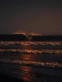 Dark elemental photograph: waves breaking on a black sand beach at ember dusk, the crest of the next swell dissolving into small glowing marks.