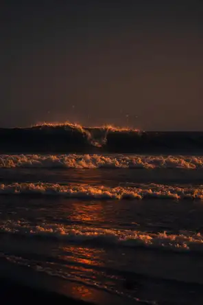 Dark elemental photograph: waves breaking on a black sand beach at ember dusk, the crest of the next swell dissolving into small glowing marks.