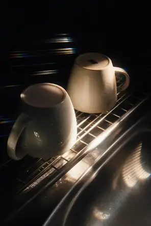 Prism-light photograph: Two mugs upturned on a wire drying rack beside a sink, one still wet, hard sun combing spectrum off the wires.