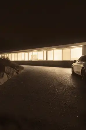 Struck-light photograph: a long modern house on a dark hillside at night, its window band lit end to end, one car parked close against it on the gravel