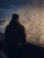 Motion photograph: a man braced and completely still at the end of a concrete sea wall at dusk as a heavy wave bursts up the wall behind him