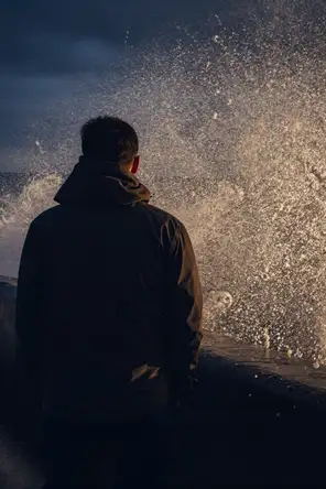 Motion photograph: a man braced and completely still at the end of a concrete sea wall at dusk as a heavy wave bursts up the wall behind him