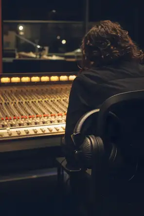 Dark cinematic photograph: A radio studio desk with a wall of faders lit warm, headphones hooked over the arm of the chair, one operator's back at the glass.