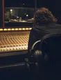 Dark cinematic photograph: A radio studio desk with a wall of faders lit warm, headphones hooked over the arm of the chair, one operator's back at the glass.