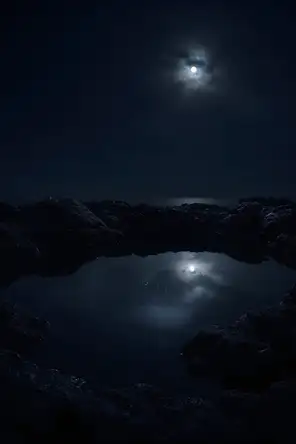 Dark elemental photograph: a still tidal pool in dark volcanic rock under moonlight, the sky's reflection in the water rendering as small glowing marks.
