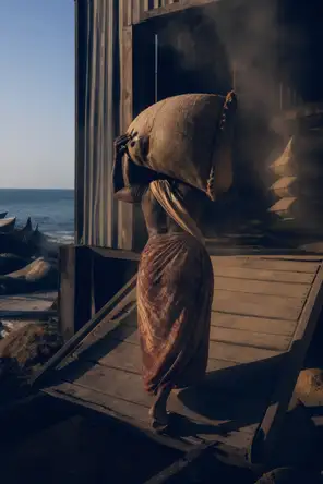 Dark cinematic photograph: One woman shouldering a full sack up a ramp into a grain store, hard low side light, dust in the air.