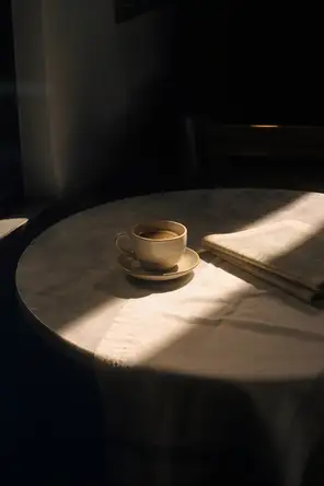 Prism-light photograph: A small cafe table set for one by a window, hard sun raking across the marble top and a folded newspaper.