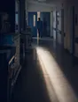 Dark cinematic photograph: A hospital ward corridor at first light, one nurse's back going in through a door, light lying along the floor.
