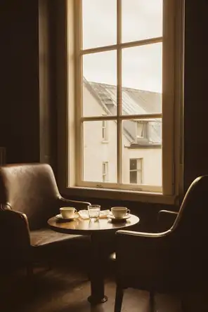 Struck-light photograph: two chairs drawn close across a low table at a tall window on an overcast afternoon, both cups used and pushed aside
