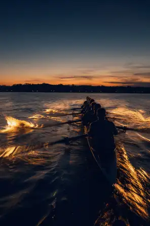 Motion photograph: a rowing eight mid-stroke on open water at first light, the blades catching and throwing water lit from behind
