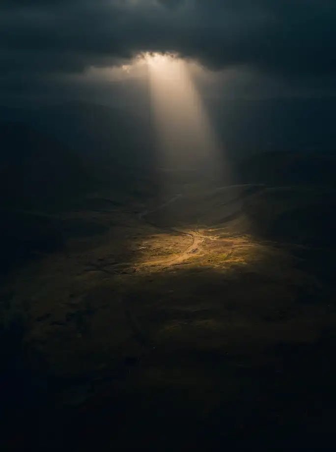Dawn aerial photograph: a shaft of first light breaking through clouds onto a single patch of dark valley.