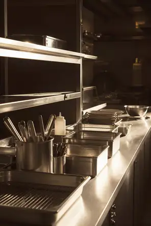 Struck-light photograph: the steel pass of a professional kitchen in the morning, wiped down and squared off before service