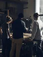 Dark cinematic photograph: Exactly three people standing around a workbench in a bright workshop, one mid-sentence with their back to us, the other two still and listening.