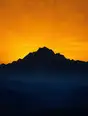 Grainy gradient illustration: A massive mountain ridge black against the first amber light of dawn, the valley below still in deep blue shadow.