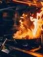 Motion photograph: a pan flaring on the burner at a professional kitchen pass, the flame climbing and steam moving through the light