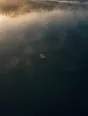 Dawn aerial photograph: a rowboat mid-lake in dawn mist, its wake trailing back toward the dock.