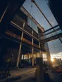Dark cinematic photograph: A huge glass panel swinging on a crane high above a half-built facade at golden hour, seen steeply from below.