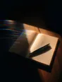 Prism-light photograph: An open blank notebook with a pen laid across it, hard sun raking across the paper grain.