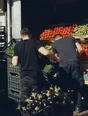 Dark cinematic photograph: A market stall at full tilt in strong morning light, crates going out fast, two traders' backs and forearms, produce stacked high.