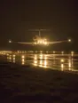 Struck-light photograph: a small aircraft on short final over a wet runway at dusk, the edge lighting running away behind it