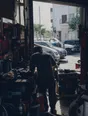 Dark cinematic photograph: A home garage turned into a working business, roller door up onto a bright street, one person's back at a crowded workbench.