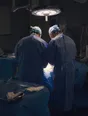 Dark cinematic photograph: Exactly two gowned figures and nobody else, seen from behind, working close together under the hard white pool of an operating lamp.