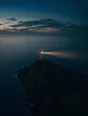 Dawn aerial photograph: a lighthouse on a dark headland at the edge of dawn, its beam still turning.