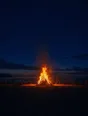 Film photograph: a bonfire blazing on a night beach, a ring of distant silhouettes around it, sparks rising into an indigo sky.