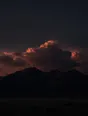 Dark elemental photograph: a towering cloud bank in deep ember light above a black ridge, its highest crown dissolving into glowing marks.