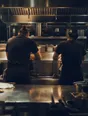 Dark cinematic photograph: A kitchen at the end of service, exactly two cooks leaning on the pass from either side, shoulders loose, warm light on wiped steel.