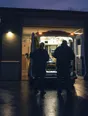 Dark cinematic photograph: An ambulance backing into a lit bay at dusk, exactly two crew waiting at the open doors.