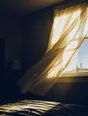 Motion photograph: a bedroom curtain thrown open to hard morning sun, dust lifting through the beam and the curtain still moving
