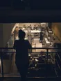 Dark cinematic photograph: One person at a mezzanine rail looking down over a hotel kitchen in full swing below.