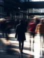 Motion photograph: a man in a good overcoat walking sharp through a bright station concourse while the crowd streams around him