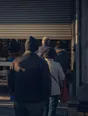 Dark cinematic photograph: A queue of backs waiting outside a small shop as the shutter goes half up at dawn.