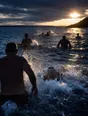 Motion photograph: open-water swimmers of different ages and builds wading and diving into a cold sea at dawn, thrown spray lit from behind