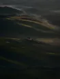Dawn aerial photograph: misted green hills at dawn with a single farmhouse, its window lit.