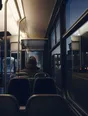 Dark cinematic photograph: A bus at first light with its interior lit, one passenger's back near the front, the dark town going past the glass.