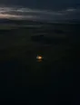 Dawn aerial photograph: a single tent glowing warm in a vast dark meadow at the first hint of dawn.