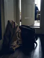 Dark cinematic photograph: A coat and a work bag set down together in a bright hallway, the door still open behind them.