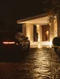 Struck-light photograph: a dark car pulling onto wet gravel under a lit portico at night, its headlights raking the columns