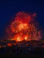 Film photograph: a fireworks finale bursting crimson and gold over town rooftops, a distant crowd watching in silhouette from a hillside.