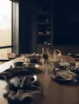 Dark cinematic photograph: A home kitchen table mid-morning, breakfast finished and pushed back, sun lying across it.